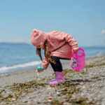 Barn i pink jakke og lilla gummistøvler, der samler skaller på stranden.